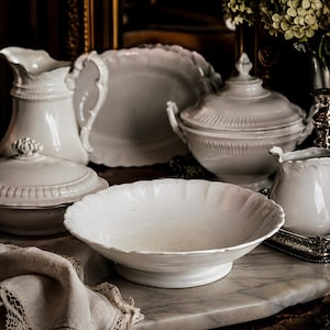 May include: A collection of white ceramic tableware including a shallow scalloped bowl, a lidded tureen, a pitcher, and a platter. The pieces feature embossed detailing and smooth glazed surfaces, arranged together to form a coordinated dining set.