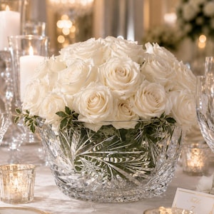 May include: A large, clear crystal bowl with intricate starburst and fan-shaped cut patterns, filled with a dense, rounded arrangement of fresh white roses and small green leaves, creating a centerpiece for a formal table setting.