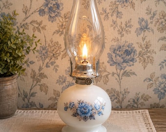 May include: A white milk glass oil lamp with a floral pattern featuring blue roses and green leaves. The lamp has a clear glass chimney, a metal burner assembly with a circular adjustment knob, and a glowing flame visible inside the glass.