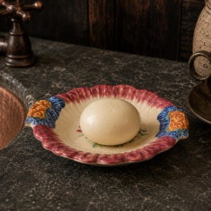 May include: A round ceramic soap dish with a scalloped, ruffled edge painted in shades of red and blue with orange floral accents. The dish features a cream-colored interior and holds a smooth, round, off-white bar of soap in the center.