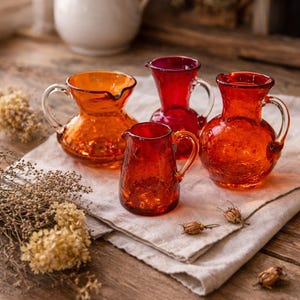 May include: Four small, colorful glass pitchers in shades of orange and red sit on a linen cloth. The pitchers have handles and textured surfaces. A white pitcher is in the background, along with dried flowers, all on a wooden table.