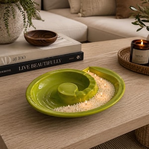 May include: A lime green ceramic decorative dish with a spiral design, filled with white beads and small lit candles. The dish sits on a light wood coffee table, alongside books, a wooden bowl, and a lit candle in a glass jar.