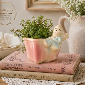 May include: A pink ceramic planter shaped like a bathtub, with a smiling seal figurine, holding a small green plant. The planter sits atop two vintage books with the titles "Child Life" and "A Guide to Garden Flowers". The image includes the text "Needlework Farmhouse".