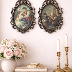 May include: Two framed oval paintings with ornate dark bronze frames hang on a white wall. Below, a bouquet of pink roses sits in a white vase, with two gold candle holders and candles to the right.