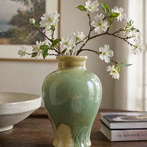 May include: A green ceramic vase with a cream-colored base holds branches of white flowers. The vase sits on a wooden table, with a bowl and books in the background. The vase has a glossy finish.