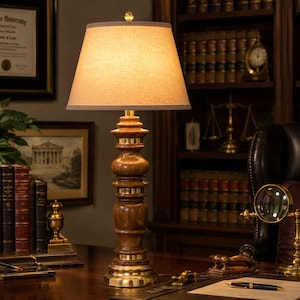 May include: A table lamp featuring a turned wood base with polished brass accents and a tapered, light-colored fabric lampshade. The lamp emits a warm glow, highlighting the wood grain and the metallic finish of the base components.