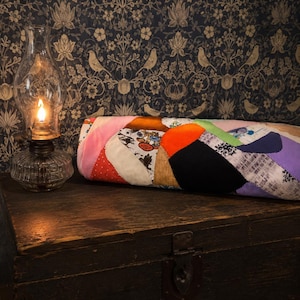 May include: A vintage-style scene featuring a lit glass oil lamp and a colorful patchwork quilt resting on a dark wooden trunk. The quilt displays various fabric patterns and colors. The background showcases a floral wallpaper.