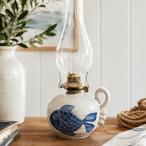 May include: A ceramic oil lamp with a rounded off-white base featuring a hand-painted blue fish design. The lamp includes a brass burner mechanism and a tall, clear glass chimney. A curved handle is attached to the side of the ceramic base.