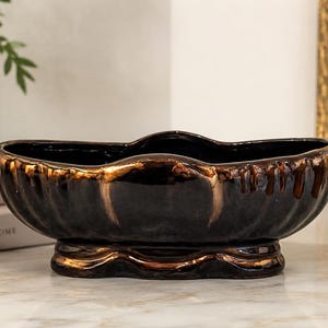 May include: An oval ceramic bowl with a glossy black finish and metallic bronze drip accents along the scalloped rim and base. The vessel features a fluted exterior texture and sits on a raised, contoured pedestal foot, showcasing a dark color palette.