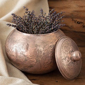 May include: A copper-colored, round container with a lid, filled with dried lavender. The container has embossed floral designs. The lid is off to the side, revealing the contents. The background includes a wooden surface and a cream-colored cloth.