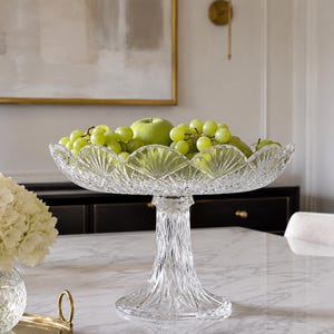 May include: A clear, cut-glass pedestal bowl featuring a scalloped rim with a fan-like pattern and a diamond-cut base. The bowl is filled with green grapes and a single green apple, showcasing intricate light-refracting geometric designs.