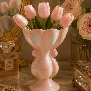 May include: A pink ceramic vase with a scalloped rim holds a bouquet of pink tulips. The vase has a bulbous shape and a pedestal base. A gold-framed mirror and a bottle of perfume are in the background.