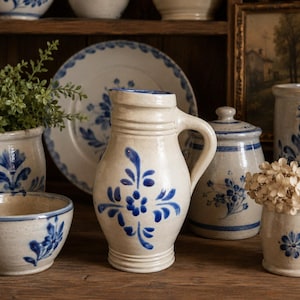 May include: A ceramic pitcher with a cream-colored glaze and hand-painted blue floral motifs. The pitcher features a rounded body, a flared rim, and a sturdy side handle. The design includes symmetrical blue petals and leaves centered on the front.