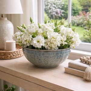May include: A large, round, blue-green ceramic bowl with a textured, embossed leaf pattern around the exterior, filled with a dense arrangement of white flowers with green buds and stems, creating a full, rounded floral display.