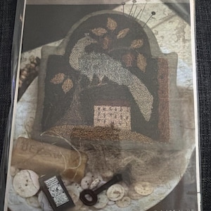 May include: A Pin Feathers design by Brenda Gervais needlework pattern in a plastic sleeve. The image shows a peacock, a house, and leaves in dark tones. Included are a small rectangular box, a metal key, and buttons. Text reads 6 1/2" X 6".