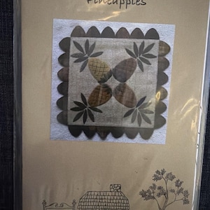May include: A craft pattern package labeled Pineapples #CCFW0005 featuring a square fabric block with four pineapple motifs in brown and tan tones, surrounded by a scalloped border. The packaging includes a line drawing of a cottage and a tree.