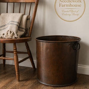 May include: A rustic, antique-style copper metal bucket with two handles, next to a wooden chair with a fringed blanket. The image includes the text "Needlework Farmhouse: Curated Objects of Enduring Beauty" in a gold circle.