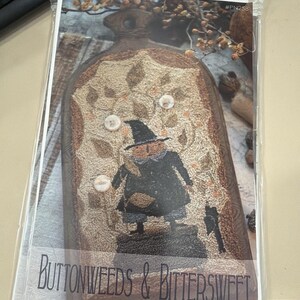 May include: A craft pattern packet titled Buttonweeds & Bittersweet by Brenda Gervais, labeled #PN258. The cover features a hooked rug design of a figure in a black coat and hat standing near a cauldron, accented with three white buttons.