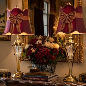 May include: A pair of table lamps featuring brass bases with clear glass accents and deep burgundy fabric shades. Each shade is adorned with a matching fabric bow. The lamps are positioned on either side of a floral arrangement and books.