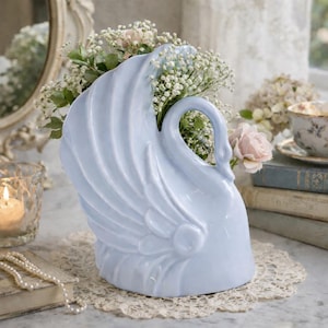 May include: A light blue ceramic swan vase filled with white and pink flowers. The swan has detailed wing and feather designs. The vase is on a lace doily, with a candle and books in the background. A vintage-style decorative piece.