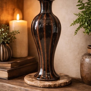 May include: A tall, dark brown ceramic vase with a glossy finish and vertical ribbed texture. The vase features a narrow neck that flares slightly at the rim and a wider, rounded body that tapers down to a circular base, standing on a stone disc.