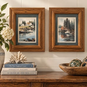 May include: Two framed watercolor prints featuring coastal scenes with houses, boats, and pine trees, each enclosed in a medium-toned wood frame with a dark blue mat. The artwork depicts calm water, rocky shorelines, and small white vessels.
