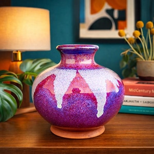 May include: A decorative ceramic vase with a wide, rounded body and a narrow neck. The vase features a vibrant color scheme of deep purple, white, and red. The vase sits on a small, round, terracotta-colored base. The background includes a lamp and books.