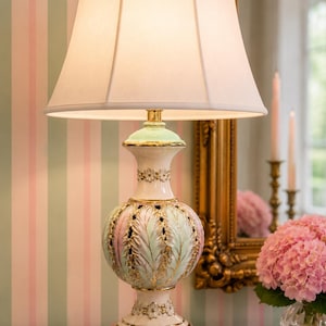 May include: A table lamp with a white fabric shade and a ceramic base featuring a pastel green and pink feather-like relief pattern with gold accents. The base sits on a square white marble plinth and includes small floral details in white and gold.