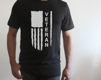 Può includere: Maglietta nera con un design a bandiera americana bianca usurata e la parola "Veteran" verticalmente sul lato destro della bandiera.