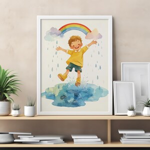 Op de afbeelding: Aquarel kunstafdruk van een kind dat in een plas springt onder een regenboog. Het kind draagt een geel shirt, een blauwe korte broek en gele laarzen. Regendruppels en wolken zijn ook afgebeeld. De print is ingelijst in een witte lijst.