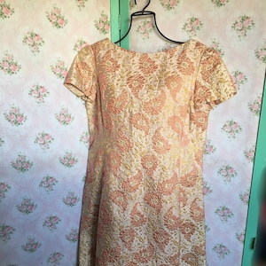 Puede incluir: Un vestido vintage con estampado floral dorado y melocotón. El vestido tiene un escote redondo y mangas cortas.