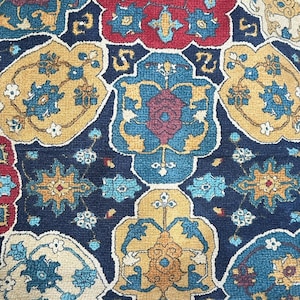 Puede incluir: Un textil rectangular con un fondo azul oscuro y un patrón repetitivo de formas florales y geométricas en rojo, amarillo y azul. El diseño recuerda a una alfombra o tapiz tradicional.