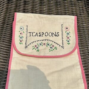 May include: A white linen tea spoon holder with pink trim and embroidered flowers. The holder has the word "TEASPOONS" embroidered in black thread.