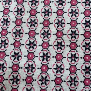 May include: Close-up of a fabric with a repeating pattern. The design features black floral shapes within pink hexagonal frames, set against a cream-colored background. The fabric has a textured appearance.