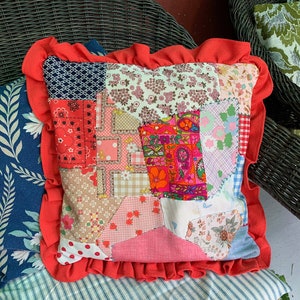 Pode incluir: Uma almofada de patchwork com uma borda vermelha com babados. A almofada é feita de vários tecidos com padrões florais e geométricos em tons de rosa, vermelho, azul e verde.