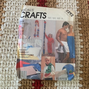Puede incluir: Un sobre de patrones McCall's Crafts vintage con varios proyectos de manualidades. La portada incluye imágenes de muñecas, un niño con una tabla de altura y una pareja con toallas. El texto incluye "Easy McCall's" y "Sick Gifts".