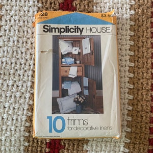 Puede incluir: Un paquete de patrones de costura Simplicity House vintage. El paquete presenta una ilustración de ropa de cama decorativa y el texto "10 adornos para ropa de cama decorativa". El paquete está sobre una superficie de ganchillo.