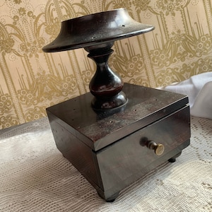 Può includere: Una scatola di legno scuro con un cassetto e un piccolo top rotondo in legno con un design decorativo. La scatola è appoggiata su un tovagliolo di pizzo bianco.