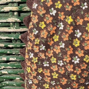 May include: Close-up of a brown fabric with a floral pattern in shades of orange, yellow, and white. The fabric has a scalloped edge and is partially visible against a green wicker background.
