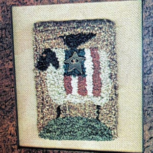May include: A framed artwork with a textured finish, depicting a sheep with an American flag design. The sheep has a black head and a star-shaped embellishment. The background is beige, and the frame is brown. A patriotic decorative item.