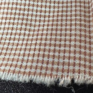 May include: Close-up of a fabric with a brown and white checkered pattern. The fabric has a frayed edge, revealing the texture of the material. The pattern is a small, repeating grid of brown and white squares.