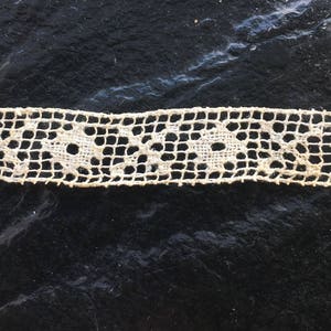May include: A close-up of a white lace trim with a delicate floral pattern. The lace is on a dark background.