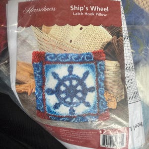 May include: A latch hook pillow kit featuring a nautical ship's wheel design. The pillow has a blue and white wheel on a white background, framed by a blue border with red accents. The finished size is 16 x 16 inches.