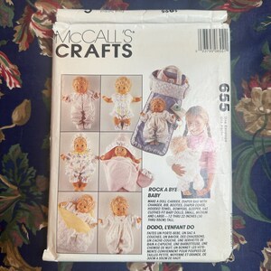 Könnte beinhalten: McCall's Crafts Schnittmuster für eine Rock-a-Bye Baby Puppentrage, Wickeltasche und Kleidung. Das Schnittmuster enthält Anleitungen für Puppen von 30 bis 55 cm Größe. Die Verpackung zeigt Bilder von Puppen.