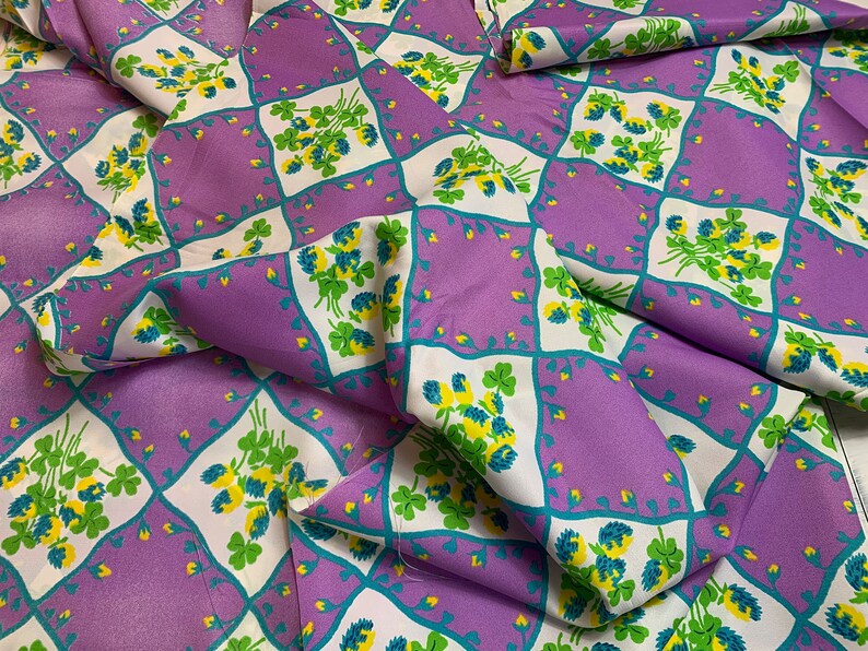 Pu&ograve; includere: Tessuto viola con un motivo a trifoglio bianco e verde. Il tessuto &egrave; piegato e presenta un motivo a rombi.
