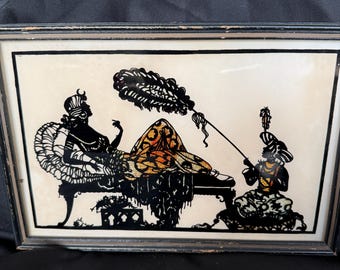 May include: Framed silhouette art depicting a reclining figure attended by another. The figures are black, with gold and orange accents on the reclining figure's clothing. The artwork is framed in a black wooden frame.