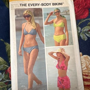 May include: A vintage Simplicity sewing pattern for a bikini, featuring three different styles. The pattern is for sizes 12-14 and is titled "The Every-Body Bikini".