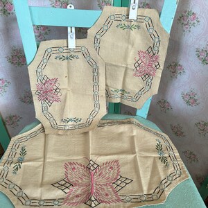 May include: Three beige fabric doilies with embroidered butterfly designs. The doilies have a black and blue border with a geometric pattern. The doilies are hanging on a light blue chair.