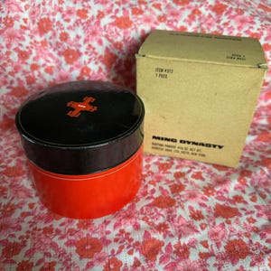 May include: A red and black round container with a black lid and a red cross design on the lid. The container is labeled "Ming Dynasty" and "Dusting Powder 4 1/2 oz Net Wt".