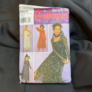 May include: Simplicity pattern 4865 for adult costumes. The pattern includes three costumes: a witch, a fairy, and a vampire. The witch costume features a black dress with a crescent moon pattern and a black witch hat. The fairy costume features a white dress with gold polka dots and a white fairy wings. The vampire costume features a red dress with a black lace overlay and a black choker.